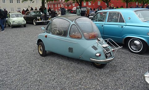 Pale blue bubble car from behind on gravel with a chrome rack, classic cars and visitors around it. Opens a larger view
