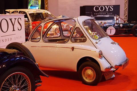 White bubble car on a red carpet beside auction signage and a dark vintage car. Opens a larger view