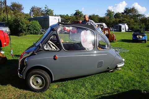 Grey bubble car with a rear luggage rack on a sunny campsite lawn, caravans behind. Opens a larger view