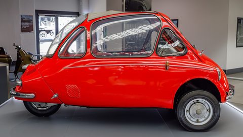 Red bubble car in side view on a grey platform, its rounded glasshouse and tail fully visible.