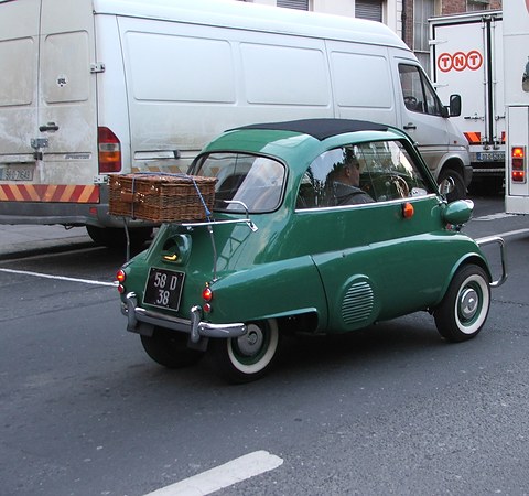 A green bubble car with whitewall tyres and a basket on its rear rack in a street, a white van behind it. Opens a larger view