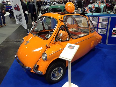 Orange bubble car with wire wheels on a blue carpet, a picnic hamper inside and an orange balloon on the roof. Opens a larger view