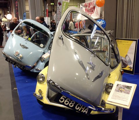Two bubble cars on a blue carpet: a cream and yellow one with its front door raised, a pale blue one behind.