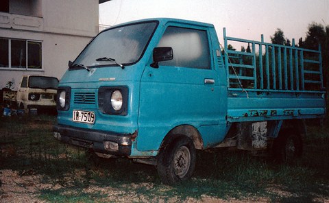 Faded blue small cab-over pick-up with a tall rack on its bed standing in a weedy yard. Opens a larger view