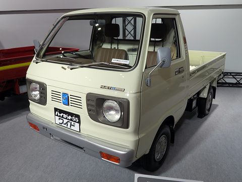 Cream small cab-over pick-up with square headlamps and a ‘55 Wide’ plate on a grey show floor. Opens a larger view