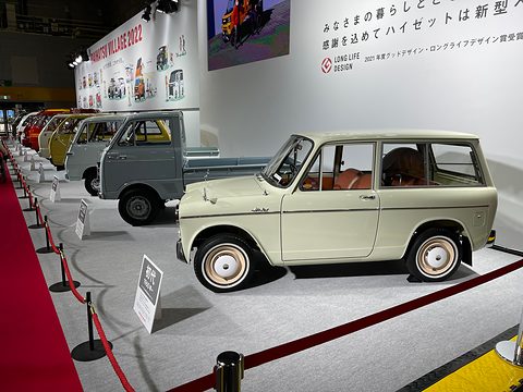 A row of small vans and trucks on a grey stand beside a red carpet, a pale green estate van nearest.