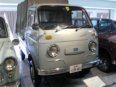 Pale blue small cab-over truck with a canvas tilt and round headlamps beside other small cars. Opens a larger view