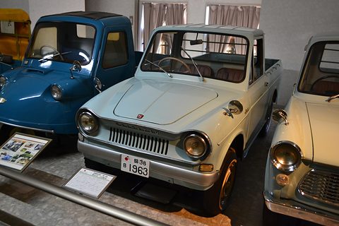 Pale blue small bonneted pick-up with a chrome grille between a blue three-wheeler and a cream pick-up. Opens a larger view
