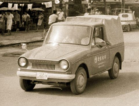 A small bonneted pick-up with a canvas cover and lettering on its door parked on a street, people with umbrellas behind.