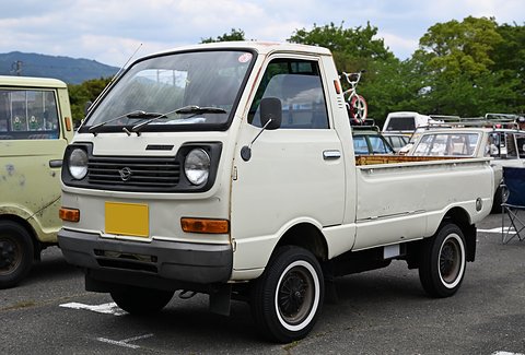 White small cab-over pick-up with black-wall tyres and a yellow plate in a car park. Opens a larger view