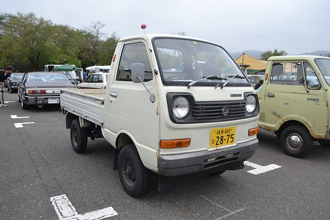 White small cab-over pick-up with a yellow plate in a car park, a green pick-up behind. Opens a larger view