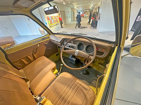 Inside a small van: tan vinyl seats, a thin steering wheel and a simple dashboard. Opens a larger view
