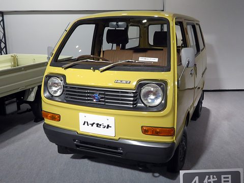 Front of a yellow small cab-over van with round headlamps and a black grille on a show floor. Opens a larger view