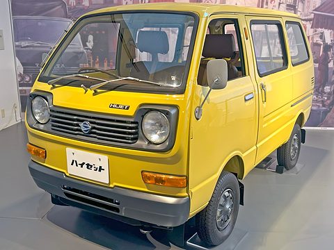 Yellow small cab-over van with round headlamps and a black grille on a grey floor. Opens a larger view