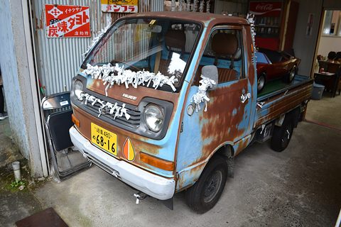 Rusty blue small cab-over pick-up with white paper decorations on its bonnet outside a shop. Opens a larger view