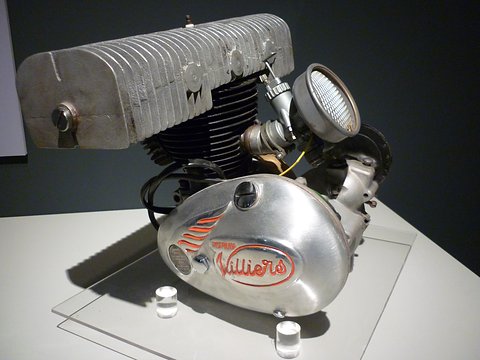 Finned silver single-cylinder engine with a fan cowl and a Villiers script on its crankcase, on a glass stand.