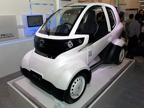 White and black tiny two-seat electric car with offset seats and open sides on a white plinth, visitors behind.