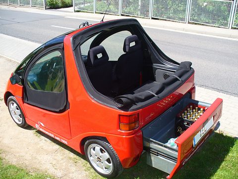 Red open two-seater with black seats and a drawer pulled out from the tail holding bottles, on paving. Opens a larger view