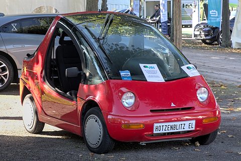 Red rounded two-seater with its door open and a HOTZENBLITZ EL plate, parked on gravel under trees. Opens a larger view