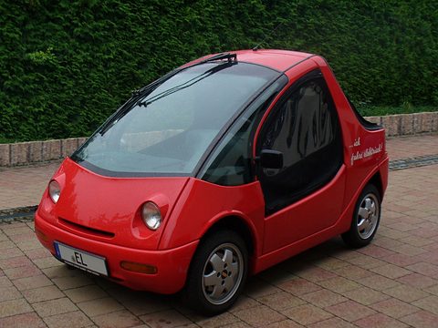 Red rounded two-seater with large round headlamps and dark side windows on brick paving before a hedge.