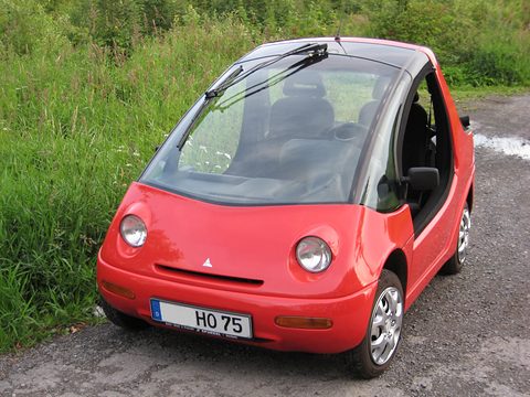 Red rounded two-seater with large round headlamps and no doors parked on gravel beside long grass. Opens a larger view
