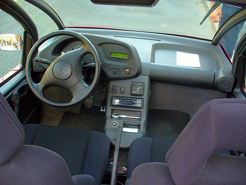 Grey dashboard with a small digital display, four-spoke steering wheel and purple-grey cloth seats. Opens a larger view