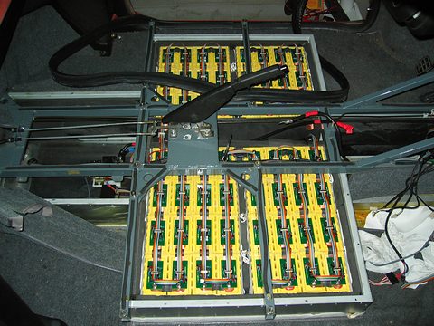 Steel tray holding rows of yellow lithium cells with green circuit boards and ribbon cables, under a lifted floor. Opens a larger view