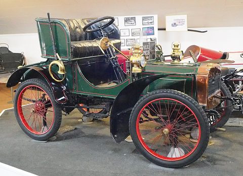 Green veteran two-seater with red wire wheels, brass lamps and a copper bonnet in a museum hall. Opens a larger view