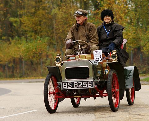 Silver veteran two-seater with red wire wheels and two people aboard on a road, autumn trees behind. Opens a larger view