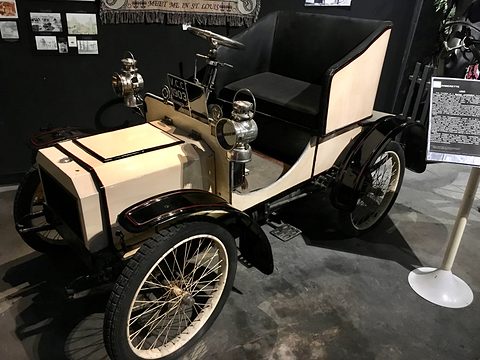 Cream veteran two-seater with black seats and wire wheels in a dark museum, a text panel beside it. Opens a larger view