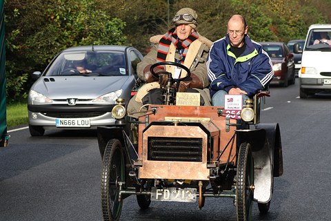 Front of a veteran car with a copper bonnet and two men aboard, modern cars following. Opens a larger view