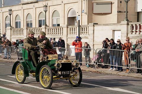 Green veteran two-seater with yellow wire wheels and two men aboard, spectators behind barriers.