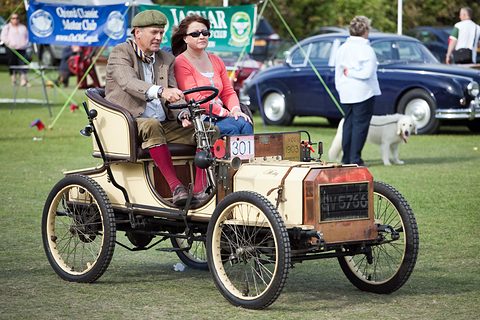 Cream veteran two-seater with a man and a woman aboard on a grass paddock, a dog and cars behind. Opens a larger view
