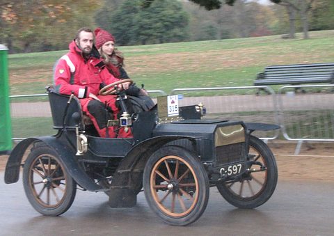 Black veteran two-seater with wooden wheels and two people in red coats on a wet road. Opens a larger view