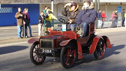 Red veteran two-seater with two men in winter coats driving past spectators on a pavement. Opens a larger view