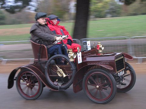 Maroon veteran two-seater with two people in coats, the background blurred by panning. Opens a larger view