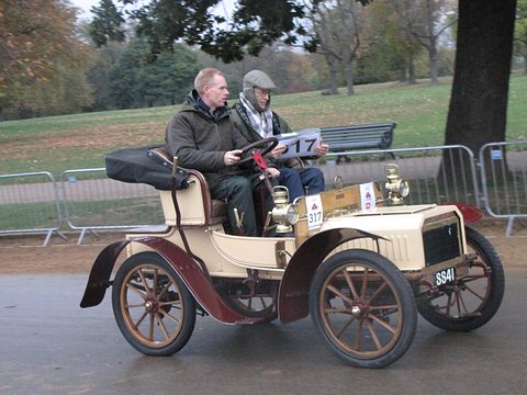 Cream veteran two-seater with maroon wings and wooden wheels carrying two men past park railings. Opens a larger view