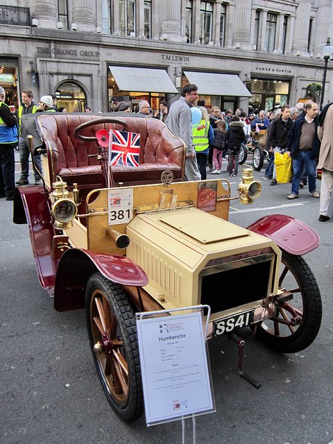 Cream veteran two-seater with maroon seats and a Union flag, an information board beside it, crowds behind. Opens a larger view