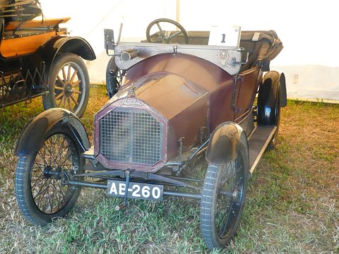 Front of a maroon small open car with an octagonal mesh radiator and a white number plate on grass. Opens a larger view