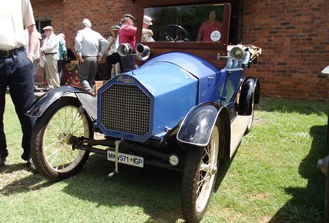 Blue small open two-seater with a mesh radiator and cream wire wheels on grass, people behind. Opens a larger view