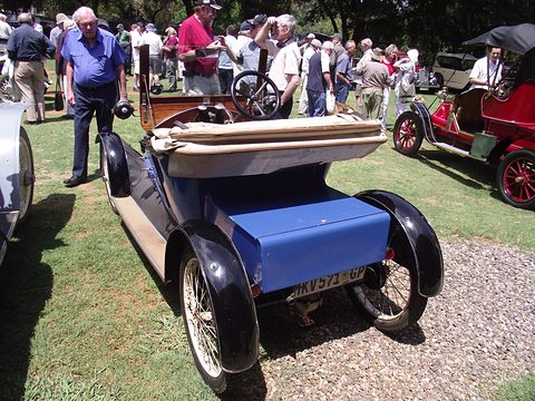 Rear of a blue small two-seater with a folded hood and black wings, a crowd behind. Opens a larger view