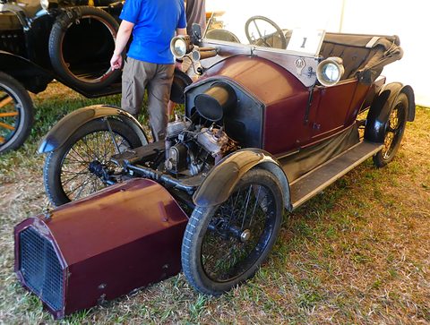 Small maroon open car with its bonnet lying on the grass, exposing a V-twin engine at the front. Opens a larger view