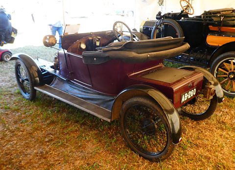 Rear of a maroon small two-seater with a folded hood and black wings in a marquee. Opens a larger view