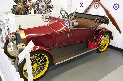 Maroon small open two-seater with yellow wire wheels beside aero engines and a propeller on display. Opens a larger view