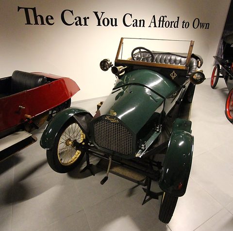 Green small open car with a mesh radiator and cream wire wheels below a wall slogan. Opens a larger view