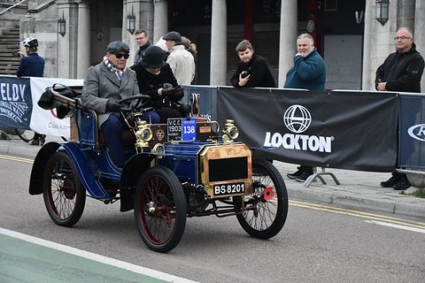 Blue veteran two-seater with red wire wheels and two men in caps, spectators behind banners. Opens a larger view