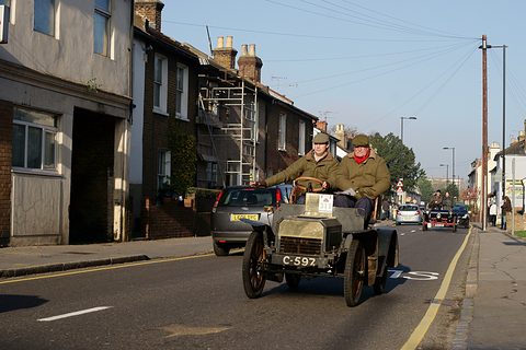 A small veteran car with two men in caps driving along a street of old houses, cars following. Opens a larger view