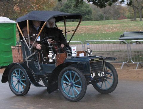 Dark veteran two-seater with a black canopy, blue wheels and a wicker basket on a wet road. Opens a larger view