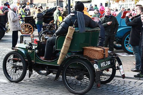 Green veteran two-seater with a wicker hamper and a rolled umbrella, its driver aboard among a crowd. Opens a larger view