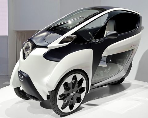 White and navy narrow three-wheeler with large alloy wheels on a white stand, seen from the front. Opens a larger view